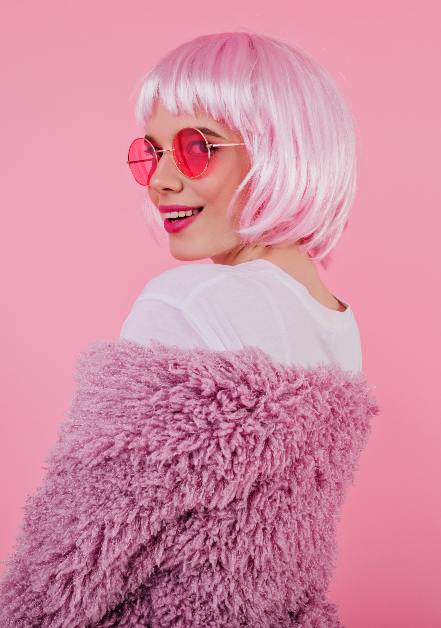 Portrait of playful girl in shiny pink peruke and sunglasses. Debonair caucasian woman looking over her shoulder with smile.